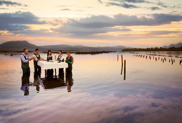 塔斯马尼亚州，歌斯湾，一群人穿着防水装围在赛菲尔菲欣纳酒店菲欣纳海洋生蚝农场的水上餐桌旁，参与生蚝品尝活动 © 澳大利亚旅游局版权所有