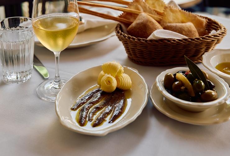 新南威尔士州，悉尼，Osteria Mucca 餐厅，凤尾鱼酱和黄油、意式面包、腌渍橄榄和一杯白葡萄酒 © Osteria Mucca 餐厅版权所有